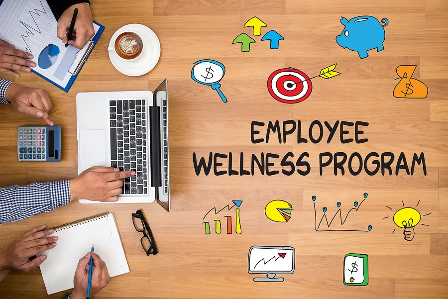 Desk with laptop, hands working on documents, coffee cup, and colorful icons representing employee wellness program elements including piggy bank, target, growth chart, and lightbulb